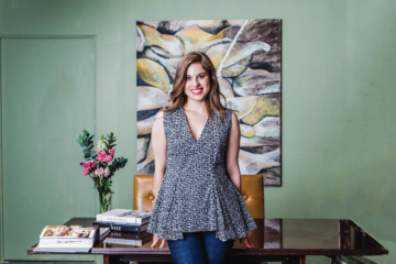 Delia Folk stands in front of a desk with a large piece of artwork behind it.
