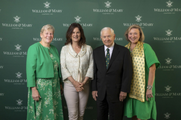 Four people pose for a photo together in front of a green wall with multiple William & Mary logos on it
